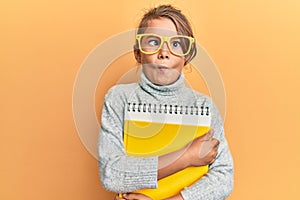 Little beautiful girl wearing glasses and holding books making fish face with mouth and squinting eyes, crazy and comical