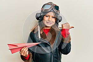 Little beautiful girl wearing pilot uniform holding paper plane pointing thumb up to the side smiling happy with open mouth