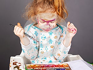 Little beautiful red-haired girl is played with paints on a dark background. the child painted his face in watercolor