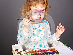 Little beautiful red-haired girl is played with paints on a dark background. the child painted his face in watercolor