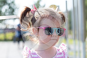 Little beautiful sad girl in sun-protective glasses with pigtails stands on the street