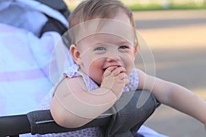 little, beautiful, smiling, cute redhead baby in a pram out-of-doors in a sleeveless shirt holding fingers in his mouth