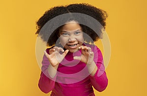 Little black girl imitating tiger, roaring and grimacing over yellow background