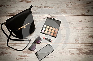 Little black ladies handbag, eye shadow, sunglasses, phone and lipstick on wooden background. Fashion concept