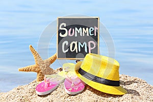 Little blackboard with text SUMMER CAMP, hat and flip flops on beach near sea