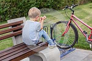 Little blond alone upset boy in casual jeans wear sitting on wooden bench in park. Lonely unhappy kid outdoors