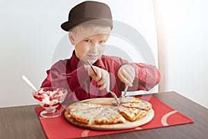 A little blond boy with blue eyes wearing red shirt and stylish cap sitting in cosy cafe eating delicious pizza and ice-cream look