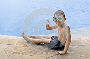 Little blond boy with goggles sits on the edge of the pool and shows thumbs up. Copy space