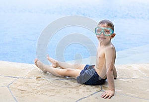 Little blond boy with goggles sits on the edge of the pool and smiles. Copy space