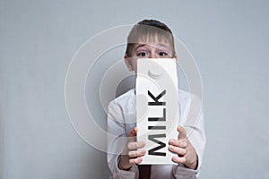 Little blond boy holds and shows a big white carton milk package. White shirt and red tie. Light background