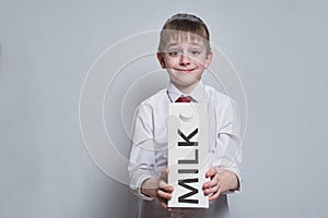 Little blond boy holds and shows a big white carton milk package. White shirt and red tie. Light background