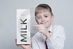 Little blond boy holds and shows the index finger on a large white carton milk package. White shirt and red tie. Light background