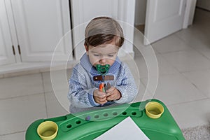 A little blond boy with a pacifier and dressed in blue, plays with some crayons on a small desk, while he is in his bedroom