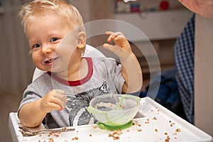 Little blond boy smiles looking at the camera because he turned over a plate of porridge and got dirty all over