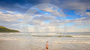 Little Blond Boy Stands in Foamy Waves of Shallow Sea on Beach