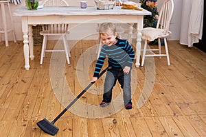 Little blond boy sweeping the floor in the kitchen. Pretty boy 3 yers old helps parents with housework.