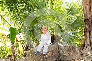 little blond boy in white clothes sitting on a stone under tropical trees