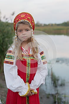 Little blond Caucasian girl in Russian national costume on blur natural landscape background