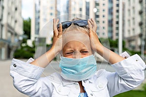Little blond caucasian girl wearing protective face mask feel stressed and unhappy screaming and crying due quarantine and covid-