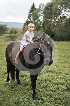 Little blond child girl in dress riding a horse. Girl on a horse walk in nature, summer field, green trees on the