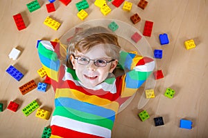 Little blond child with glasses playing with lots of colorful plastic blocks indoor. Kid boy wearing colorful shirt and