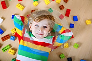 Little blond child playing with lots of colorful plastic blocks indoor. Kid boy wearing colorful shirt and having fun