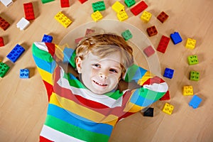 Little blond child playing with lots of colorful plastic blocks indoor. Kid boy wearing colorful shirt and having fun