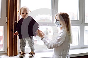 Little blond girl baby standing on windowsill near childrens doctor wearing white uniform at home, holding patient hand.