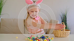 Little blond girl in bunny ears Looks surprised and pick up multicolored egg in basket. Easter egg hunt.