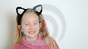 A little blond girl in cats ears headband is actively telling something and looking at the camera. The girl sings or