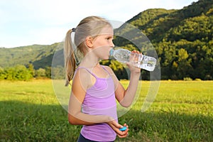 Little blond girl doing fitness exercises in the park and drinking a water
