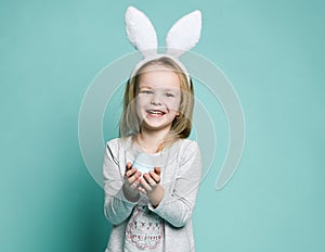 Little blond girl in gray cosy home dress and decorative fur ears standing, holding pastel blue Easter egg in hands