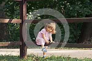 Little blond girl in pink dress climbing over the wooden fence in summer day on the forest background. Vocation in the village.