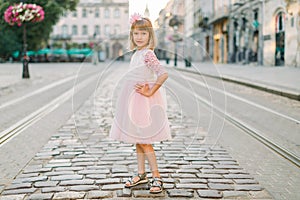 Little blond girl in pink fashionable dress standing on the pavement street road on the background of old European city