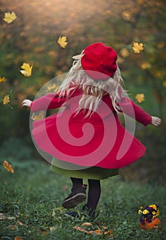 A little blond girl in a red coat and beret is spinning in yellow leaves in the autumn forest. Autumn photography.