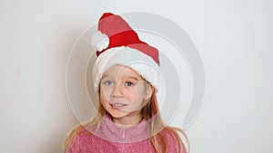 A little blond girl in red Santa Claus hat is actively telling something and looking at the camera. The girl sings or