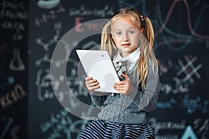 Little blond Girl in school uniform holding white tablet pc in chemistry class. Chalkboard with school formulas background