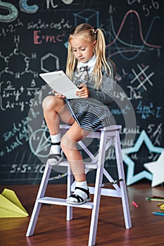 Little blond Girl in school uniform holding white tablet pc in chemistry class. Chalkboard with school formulas background