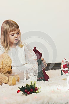 Little blond girl sitting on the floor, playing with her teddy bear and little Santa Claus Christmas toy