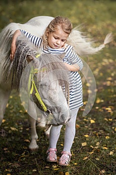 Little blond girl is standing near the little horse pony and hugging it