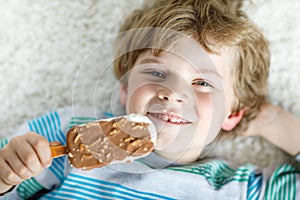 Little blond kid boy with curly hairs eating ice cream popsicle with chocolate at home