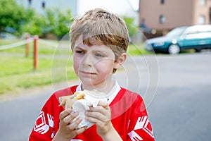 Little blond kid boy eating hot dog after playing soccer.