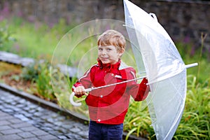 Little blond kid boy walking with big umbrella outdoors on rainy day. Portrait of cute preschool child having fun wear