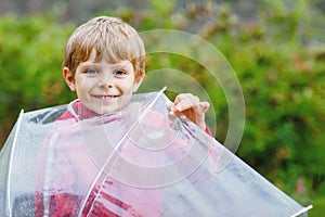 Little blond kid boy walking with big umbrella outdoors on rainy day. Portrait of cute preschool child having fun wear