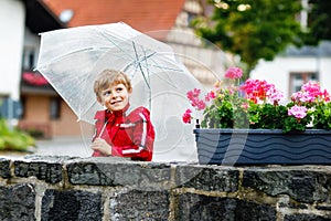 Little blond kid boy walking with big umbrella outdoors on rainy day. Portrait of cute preschool child having fun wear