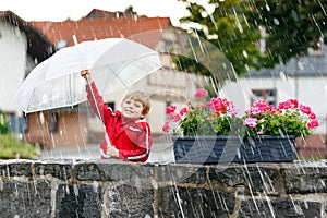Little blond kid boy walking with big umbrella outdoors on rainy day. Portrait of cute preschool child having fun wear