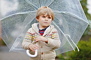 Little blond kid boy walking with big umbrella outdoors on rainy day. Portrait of cute preschool child having fun wear