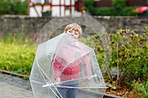 Little blond kid boy walking with big umbrella outdoors on rainy day. Portrait of cute preschool child having fun wear colorful