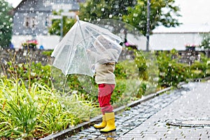 Little blond kid boy walking with big umbrella outdoors on rainy day. Preschool child having fun and wearing colorful