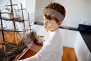 Little blond school kid boy playing with sailing ship model indoors. Excited child with yacht having fun after school at